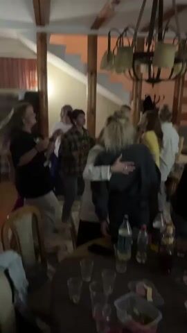 The video shows a group of women dancing and socializing indoors. The environment appears to be a room with wooden beams on the ceiling and a staircase. There is a chandelier hanging from the ceiling. In the foreground, a table is visible with several plastic cups, bottles of drinks, and some food items. The women are dressed casually and appear to be enjoying themselves. Some are dancing, others are talking and embracing. One woman is wearing a white blazer, and another is wearing a plaid shirt. The lighting is dim, suggesting it may be evening.