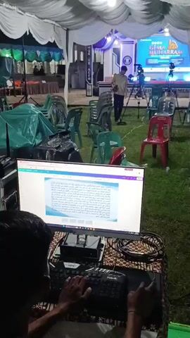 The video shows an outdoor event setup at night, likely under a large tent with white draping. In the foreground, a person is operating a computer with a keyboard and mouse, seemingly controlling some aspect of the event's technical production. The computer screen displays text, possibly a script or lyrics. Behind this workstation, there are several monitors displaying various technical data, including a grid layout that suggests video switching or monitoring. Equipment racks with electronic components and cables are visible. In the midground, an empty seating area with chairs (some green, some red) is arranged on the grass. Further back, a stage is set up with a large screen displaying text and graphics, and several people are working behind it, including someone operating a camera on a tripod. The overall atmosphere is one of event preparation or live production.