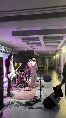 The video shows a band performing on a stage. A singer in a light-colored suit is at the microphone, and a guitarist is to his left. A drummer is behind them. The stage has a large screen with colorful graphics. The lighting is purple. In the foreground, there are speaker cabinets and musical equipment. The second part of the video shows a few people dancing in a dimly lit room, with tables and chairs in the background.