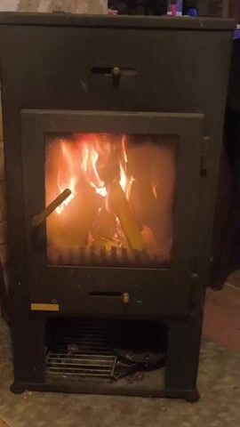 A video shows a close-up of a black wood-burning stove with flames visible through the glass door. The fire is actively burning, with logs and embers within the firebox. The stove has a metallic handle on the left side of the door, and two small knobs on the front, one above the door and one below. At the bottom of the stove, there is a metal grate. The surrounding environment appears to be indoors, with a brick wall visible on the left and a dark background on the right. The lighting is dim, with the fire being the primary light source.