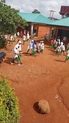 The video shows a group of children, primarily girls, in a courtyard outside a school building. The children are dressed in uniforms, predominantly white with green accents, and many wear headscarves. They appear to be moving about the courtyard, some walking, others possibly congregating. The school building has a light green roof and brick walls. The ground of the courtyard is made of reddish-brown earth. In the foreground, there is lush green foliage. A motorcycle is parked to the left of the frame. The sky is partly cloudy, suggesting daytime. The overall impression is of a school environment, likely during a break or transition period. The children's expressions are not clearly discernible, but their movement suggests general activity rather than distress. The scene is set in Kagarko South, Nigeria. There is no visible text in the video.