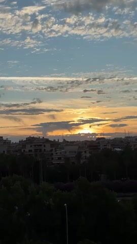 The video shows a time-lapse of a sunset over a city. The sun is visible in the center of the frame, casting a warm glow over the buildings and trees. The sky is filled with clouds, which change color from orange and pink to blue and white as the sun sets. The city appears to be a mix of residential and commercial buildings, with a lot of greenery, including palm trees and other foliage. There are no people or animals visible in the video. The video is shot from a high vantage point, likely a balcony or rooftop, offering a panoramic view of the cityscape.