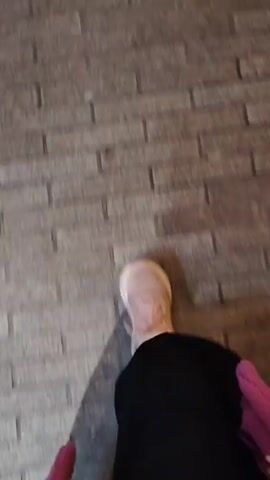 A person wearing a light-colored shoe, black pants, and a red and pink patterned top walks across a brick walkway. The video is shot from a first-person perspective, focusing on the lower body and the ground. The walkway consists of horizontally laid bricks in varying shades of gray. The camera is unsteady, shaking slightly with each step.