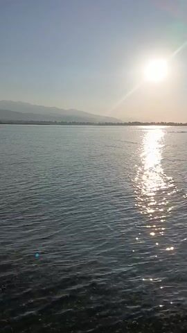 The video captures a wide expanse of water under a bright sun. The sun is high in the sky, casting a strong glare on the water's surface, creating a shimmering reflection. In the distance, a faint, hazy outline of mountains or land can be seen on the horizon. The water itself appears to be a deep blue or gray, with small waves rippling across its surface. In the middle ground, a red object, possibly a flag or a kite, is visible flying above the water. The overall scene is one of natural beauty, with the sun and water dominating the frame.