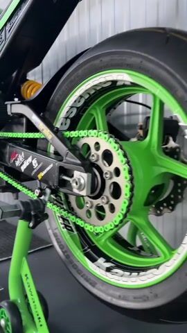 This is a close-up video of a custom Kawasaki motorcycle. The camera pans across the motorcycle, focusing on various details. In the first shot, we see the rear wheel, chain, and swingarm. The chain is bright green, and the wheel rim is also green with white decals. The motorcycle is on a green stand. The camera then moves to show the engine and frame, highlighting the black and green color scheme. We can see the exhaust system, air filters, and various mechanical components. The front of the motorcycle is shown next, with the headlight, front forks, and front wheel. The tire has green and white branding. Throughout the video, the motorcycle appears to be in a workshop or showroom environment, with a corrugated metal wall in the background. The video is well-lit, and the details of the motorcycle are clearly visible.