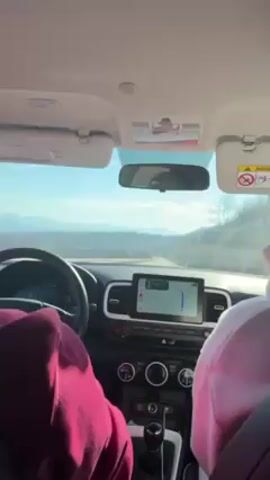 The view from inside a car shows the driver and front-seat passenger, both wearing hooded sweatshirts. The car is driving on a paved road through a rocky, hilly landscape under a clear blue sky. A navigation system is displayed on the car's dashboard screen. Mountains are visible in the distance. The car's interior is visible, including the steering wheel, dashboard, and sun visors. The car appears to be traveling during the daytime.