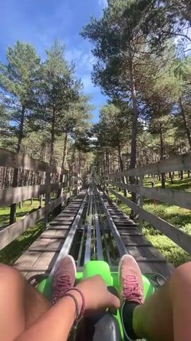 This is a first-person perspective video taken from a bobsled ride on a sunny day. The bobsled is green and is moving along a track made of metal rails and wooden planks. The track is surrounded by a forest of tall pine trees. The rider's legs and feet are visible in the foreground, wearing pink sneakers. The rider's hands are on the controls of the bobsled. The sky is blue with some white clouds. The camera is tilted upwards, looking ahead along the track. The rider's left hand is briefly raised and moved side to side.