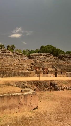 The video shows a wide shot of an ancient Roman amphitheater. The amphitheater is made of stone and has tiered seating that surrounds a central arena. The arena floor is covered in dirt and dry grass. There are several people visible in the distance, walking around the amphitheater and exploring the ruins. The sky is cloudy and hazy, suggesting a warm day. The overall impression is one of historical significance and decay.
