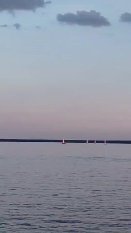 A wide shot captures a serene lake at dusk. The sky is a gradient of soft blues and pinks, with a few scattered clouds. The water is calm, reflecting the muted colors of the sky. In the distance, a line of sailboats, their white sails catching the last of the light, are spread across the horizon. Closer to the foreground, a whimsical swan-shaped paddle boat glides across the water. The overall atmosphere is peaceful and tranquil.