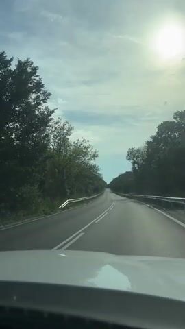 A first-person view from a vehicle driving on a two-lane road. The road has a solid white line on the right, and a double solid white line on the left, indicating no passing. Trees line both sides of the road, forming a green tunnel. The sky above is mostly clear with some scattered white clouds. The sun is visible in the upper right corner of the frame, causing a glare. The vehicle's dashboard is visible at the bottom of the frame.