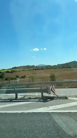 The video captures a roadside scene with a bright blue sky and a few white clouds. In the foreground, a guardrail separates a paved surface from a grassy area. A road sign with blue text in a foreign language is visible on a pole, along with other traffic signs. Beyond the guardrail, a landscape of rolling hills covered in dry grass and scattered green trees unfolds under the sunlight. Some patches of cultivated fields are also visible. The camera appears to be moving, suggesting a view from a vehicle.