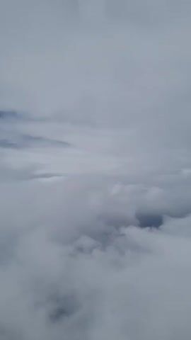 The video shows a view of thick, low-hanging grey clouds, likely taken from an elevated position, possibly an airplane. The clouds obscure the ground below, creating a uniform, overcast scene. The video remains static with no movement of the camera. There is also unintelligible speech in the background.