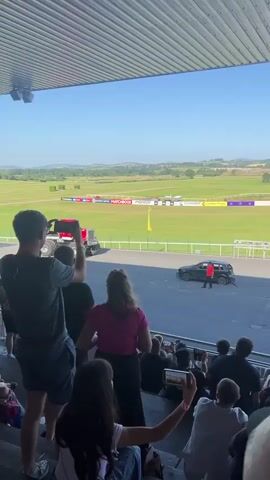 The video shows an outdoor event with spectators seated on tiered bleachers, facing a wide open field with a track. A large red monster truck is positioned on the track, and a smaller dark-colored vehicle is in front of it. A person in a red shirt is standing near the dark-colored vehicle. The field behind the track appears to be a grassy area with some signage. The sky is clear and blue, suggesting a sunny day. People in the foreground are holding up phones, presumably to record the event. The overall atmosphere appears to be that of a live show or demonstration.