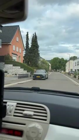 The video is filmed from inside a car, looking forward through the windshield. A smartphone is mounted on the dashboard, displaying a navigation app. The app shows a route with turns and estimated travel time. The car is driving on a paved road lined with houses and trees. The sky is cloudy. The car's radio display shows "102.60 MHZ".