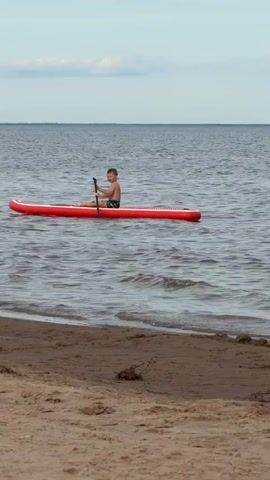 A young boy is on a red paddleboard in the water near a sandy beach. He is wearing swim trunks and holding a paddle. The water is choppy. The sky is overcast.