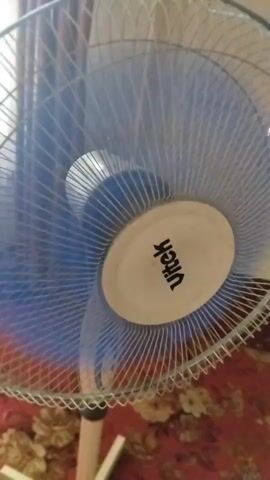 The video is a close-up shot of a Vitek brand oscillating fan. The fan blades are blue and are blurred due to their rapid rotation. The fan's central housing is white with the Vitek logo printed in black. The fan is set against a backdrop of floral patterned carpet and curtains, suggesting an indoor domestic setting. The lighting is somewhat dim, with the light source appearing to be from a window. The fan is likely in operation, as indicated by the movement of the blades.
