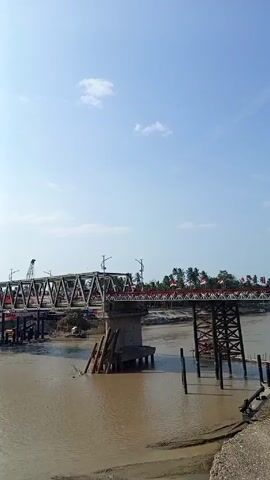 The video shows a bridge over a muddy river on a sunny day. The bridge is decorated with Indonesian flags. A crane is visible on one side of the bridge. There's some construction material and construction debris along the river bank. A white van is seen driving on the bridge.