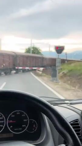 The video shows a view from inside a vehicle looking towards a railway crossing. A long freight train with many open-top brown rail cars is passing. The railway crossing gates are down, indicated by the red and white striped barriers blocking the road. A red flashing light on the signal post confirms the active crossing. Other vehicles are stopped behind the crossing gates. The time of day appears to be late afternoon or early evening, with a cloudy sky and the sun setting behind distant hills. The dashboard of the vehicle is visible in the foreground, with a speedometer indicating the vehicle is stationary. The overall environment is rural, with vegetation and trees visible on the sides of the road and railway tracks.