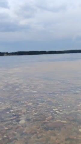This video shows a low-angle view of a body of water, likely a lake or reservoir, on a cloudy day. The foreground is dominated by a rocky, pebbly bottom visible through clear, shallow water. In the distance, a tree line forms a dark horizon.

The activity taking place is swimming. A person can be seen in the water in the middle ground, appearing to be swimming or wading. The sky is overcast with grey clouds, suggesting it might be late afternoon or an otherwise cloudy day. The overall mood is calm and peaceful, with the gentle ripples on the water adding to the serene atmosphere. The location is identified as the "Klyazminskoye Reservoir Tourist Resort" in Russia, indicating a recreational area. There is no visible text in the video.