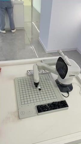 The video shows a white robotic arm playing a game of Go on a gray gridded board. The robot picks up a white stone from a tray and places it on the board next to a black stone. A person wearing jeans and white shoes is standing in the background, seen through a glass partition. The robot has a black screen displaying information and a robotic arm with a gripper. The game board has white and black stones in separate trays on either side. The table is white, and the floor is light gray with shiny lines. The setting appears to be a modern, clean environment.