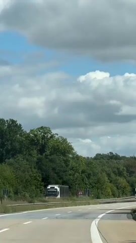 The video shows a view from inside a moving vehicle on a highway. The highway has multiple lanes and is surrounded by lush green trees. There are several vehicles on the road, including cars and trucks. The sky is partly cloudy with bright blue patches. The road is paved and has white lane markings. There are also some traffic signs visible along the roadside.