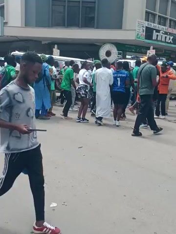 The video shows a group of people walking down a city street. Most people in the group are wearing green shirts and pants, but some are wearing orange and blue shirts. One person is wearing black Adidas sweatpants with a gray T-shirt and is holding a cell phone. The street is paved and there is a building on the left side of the video. There are signs posted on the building. A person rides by the group on a motorcycle.