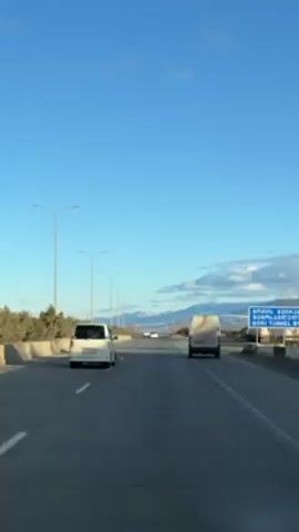 The video shows a car driving on a highway. The sky is blue and there are mountains in the distance. A white van and a white car are visible on the road. Streetlights line the road. There is a blue sign on the right side of the road with writing in a different alphabet. The camera is positioned inside a car.