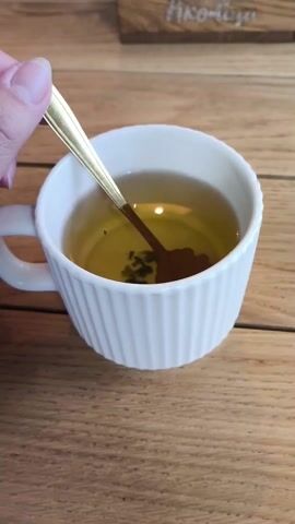 A hand is holding a gold-colored flower-shaped spoon over a cup of tea on a wooden table. The cup is white with vertical ridges and contains a yellow-green tea with some herbs floating in it. Honey is dripping from the spoon into the cup. In the background, a wooden object with writing is partially visible.