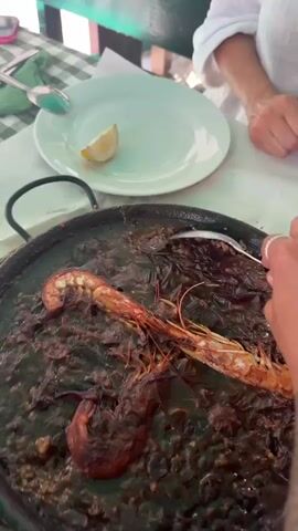 A close-up shot focuses on a dark, wet paella dish with large shrimp. A hand holding a spoon is seen stirring the rice and scooping it onto a light green plate with a lemon wedge. In the background, two individuals are partially visible, seated at a table covered with a white tablecloth.