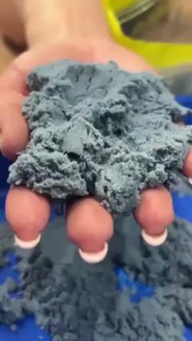 The video shows a close-up of a hand holding a clump of blue-gray kinetic sand. The sand is textured, looking somewhat like a dense, crumbly cloud. The hand has manicured nails with white tips. Some sand is falling between the fingers and onto more sand below, presumably in a container. The background is slightly out of focus, but shows a blue surface and a hint of a yellow object.