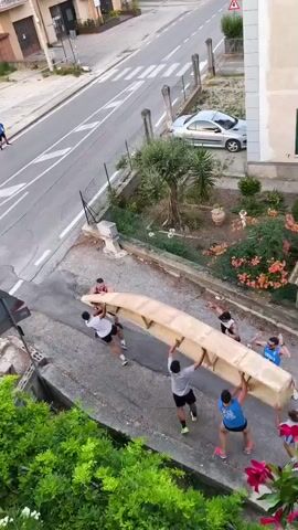 Six people are carrying a long, light-colored wooden object that resembles a coffin up a sloped path. The object is being held horizontally above their heads. There are buildings and a street in the background, with a few cars and bicycles. The people are wearing casual clothing. The lighting suggests it is daytime.