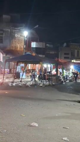 At night, a street with small market stalls lit by bright lights is visible. The street is littered with trash, mostly plastic bags. Several motorcycles are parked, and others pass by. A few people are visible around the stalls, some appearing to be vendors. The buildings in the background are old and varied in style. A street light illuminates the scene.