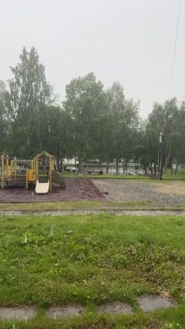 The video shows an outdoor playground during a rain shower. The playground equipment is visible on the left side of the frame, including a slide and a climbing structure. The ground around the playground is wet, indicating that it is raining. Trees and benches are visible in the background. The weather appears to be overcast and the rain is falling steadily. The scene suggests a quiet and somewhat melancholic atmosphere due to the weather. There is no visible text in the video. The location is Kandalaksha, Russia.