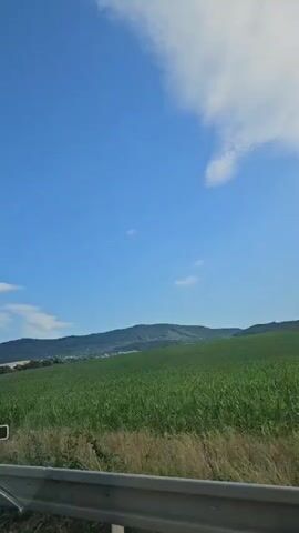 This is a video of a landscape with green fields and hills under a blue sky with white clouds. The camera is moving, possibly from a vehicle, as indicated by the presence of a blue metal railing or barrier in the foreground. The focus is on the natural scenery, with no people or significant actions visible.