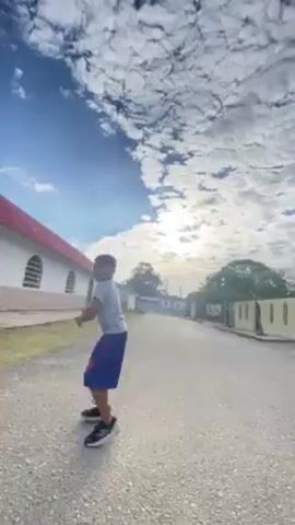 A low-angle shot in a sunny outdoor setting shows a boy throwing a football on a paved street. The boy is wearing a light-colored t-shirt, dark blue shorts, and black running shoes. He throws the football with his right arm, then walks towards the camera, giving a thumbs up. The camera then pans up to show a vibrant blue sky filled with patchy white clouds. The background features a white building with a red roof on the left side of the frame, and residential houses and trees lining the street. The overall atmosphere suggests a sunny, quiet neighborhood.