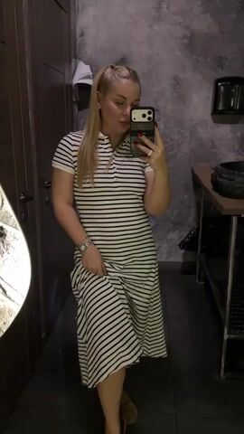 A fair-skinned woman with blonde hair in a high ponytail stands indoors, posing for a mirror selfie. She wears a short-sleeved, collared midi dress with horizontal white and dark blue stripes. She holds a dark smartphone with a patterned case in her right hand, displaying red nail polish and a ring, while looking at the phone screen. Her left hand, adorned with a silver-colored bracelet, gently holds the skirt of her dress. The background on the left features a dark wooden door with a silver handle and a white cloth hanging, while the right side reveals a textured dark gray wall, a dark wooden counter with a black bowl-shaped sink, and a black metal shelving unit below.