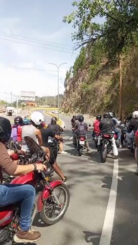 This video clip shows a group of motorcyclists riding on a road in San Antonio de Los Altos, Venezuela. The time of day appears to be daytime, with bright sunlight, suggesting fair weather. The road is paved and has lane markings. On the left side of the road, there are palm trees and buildings, including what looks like a Mercedes-Benz dealership with its distinctive logo. On the right side of the road, there is a rocky, tree-covered hillside.

The motorcyclists are all wearing helmets for safety. They are riding in a procession, with some in the foreground and others further down the road. The motorcycles are various models and colors. Some riders have backpacks, and one motorcycle in the foreground has a red cargo box with black netting. The overall mood seems to be one of a leisurely ride or a group outing. The license plate of the motorcycle in the far right foreground is partially visible and reads "AC436GL".