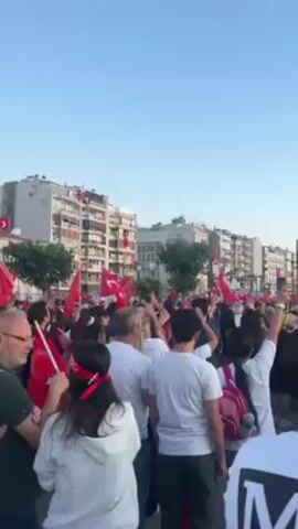Here is a concise description of the video:

The video shows a crowd of people gathered in a town square. Many are holding and waving Turkish flags. Some people are wearing white shirts, some are wearing red headbands. The environment shows multiple buildings, trees, and light posts. There is a light blue sky above.
