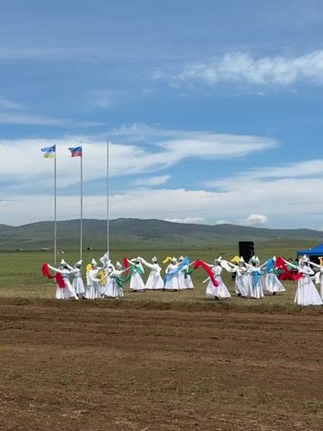 A group of performers wearing white costumes with colorful sashes are dancing in an open field. They are arranged in a semi-circle formation and appear to be engaged in a traditional dance. In the background, there are flags flying on poles, a stage with a banner that reads "АЛТАРГАНА 2020", and a cluster of tents. The environment is a grassy field with some dirt in the foreground, and hills can be seen in the distance under a partly cloudy blue sky. The overall scene suggests an outdoor cultural event or festival.