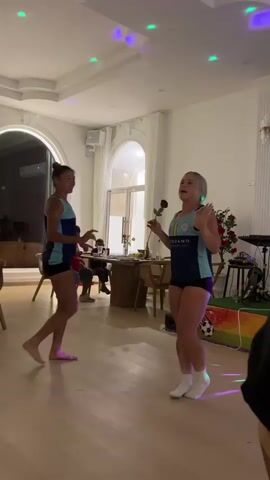 Two women in matching athletic wear are dancing in a well-lit room. The woman on the left has dark hair pulled back in a bun and is barefoot. The woman on the right has blonde hair and is wearing white socks. She is holding a red rose in her right hand. Colorful lights are projected onto the ceiling. In the background, there is a table with chairs, a person sitting at the table, and a musical instrument. The floor is light-colored wood.