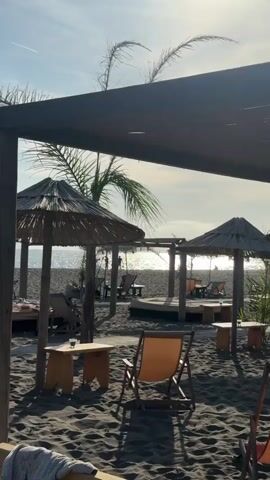The scene is a beachside setting on a sunny day, capturing a relaxed and tranquil atmosphere. The foreground features several wooden chairs and small tables arranged on the dark sand. Straw umbrellas are dotted around, offering shade to seating areas. In the background, the ocean glistens under the sun, with people visible enjoying the beach. An airplane trail can be seen in the clear, blue sky. The video is shot from under a shaded structure, providing a framed view of the beach scene.