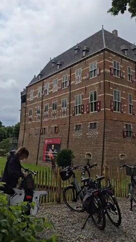 The video shows a brick building with multiple windows and red shutters, possibly a historical structure or castle. Several bicycles are parked in front of it, some with bags attached. A person is standing near a bicycle on the left side of the frame. The ground is covered with gravel, and there's a wooden fence. Trees and foliage are visible, suggesting an outdoor setting. The sky appears overcast.