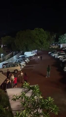 The video shows a dimly lit outdoor parking lot at night. Several cars are parked in rows, including a white van, several sedans, and an SUV. A few people are scattered throughout the parking lot. On the left side, a group of three women and two men are gathered near a white SUV, with one woman appearing to be showing something on her phone to the others. In the middle ground, two men are walking away from the camera. To the right, a woman in a green jacket stands facing away from the camera. In the foreground, a red car is prominently parked, with a person standing next to it. In the background, trees and buildings are visible, with some lights illuminating the area. The ground appears to be dirt or gravel.