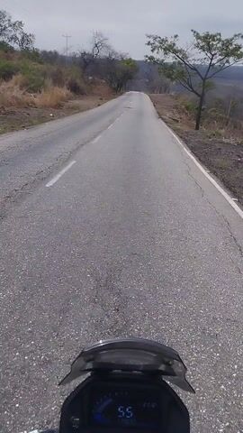 A motorcycle is moving along a paved road. The speedometer reads 55. There is dry, hilly terrain visible to the side of the road. The sky is overcast. The landscape appears to be arid, with mostly bare trees and dry vegetation. There are power lines visible in the distance.