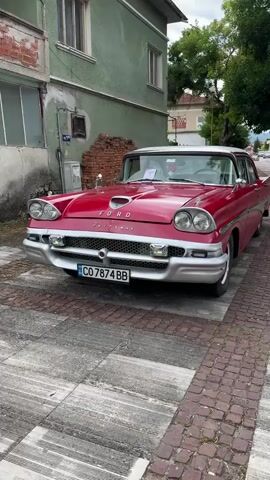 The video captures a vintage red Ford Fairlane parked on a cobblestone street. The car features a white roof, chrome accents, and a prominent grille. The background shows a weathered green building with windows and a brick wall section. Trees and a cloudy sky are visible above. A license plate with the alphanumeric characters "CO 7874 BB" is affixed to the front of the car. The camera pans slightly to the right, offering a three-quarter view of the vehicle. No people are visible in the frame.