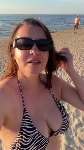 This is a selfie-style video of a woman at the beach. She is wearing a black and white zebra-print bikini top and black sunglasses. Her brown hair is loose and appears to be blowing in the wind. The woman is looking at the camera and her mouth is slightly open, as if she is speaking. The background shows a sandy beach with a few other people in the distance. There are also some beach umbrellas and tents, suggesting a developed beach area. The lighting indicates it is either early morning or late afternoon, with the sun casting a warm glow. The overall mood appears relaxed and summery. The location is identified as Jūrmala, Latvia.