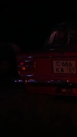 The video shows the rear of a red car at night. The car has a license plate that reads "C 666 KA 01". In the background, there are several bright white spotlights shining upwards, and a yellow structure with lights on it. A silhouetted person is standing near the car, facing away from the camera. The scene appears to be outdoors, possibly at an event or performance.