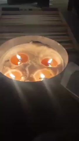 The video shows an indoor space. In the first shot, a large candle with multiple wicks sits on a table with a backgammon board. As the camera zooms out, a person is seen sitting on a couch near the table, which is lit by the large candle. Another grouping of candles is placed near a painting of a person. Other paintings hang on the wall. A chair and a glimpse of people are visible in the background, outside of the room.