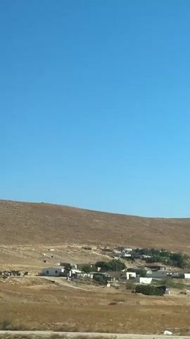 The video shows a wide shot of a desert landscape under a clear blue sky. A low, brown, dry hill dominates the midground. In the foreground, a small settlement of white buildings is visible, interspersed with sparse greenery. The ground is arid and beige, with patches of scrub vegetation. The camera appears to be moving slightly, possibly from a passing vehicle.