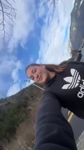 A woman in a black Adidas hoodie takes a selfie outdoors. Bare tree branches and a blue sky with clouds are visible above her.  A green, wooded hill slopes in the background. She is smiling and holding her phone at an upward angle.